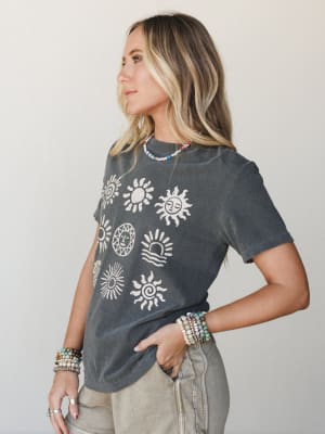 A washed charcoal graphic tee printed with a collection of illustrated sun motifs is styled casually with sage denim and layered beaded jewelry — effortless and a little celestial.