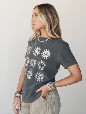 A washed charcoal graphic tee printed with a collection of illustrated sun motifs is styled casually with sage denim and layered beaded jewelry — effortless and a little celestial.