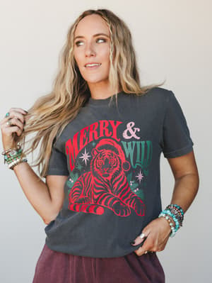 A washed charcoal graphic tee with a retro 'Merry & Wild' tiger-in-a-Santa-hat print, styled with stacked boho bracelets and a dusty mauve skirt — the kind of holiday outfit that skips the ugly sweater and still wins the party.