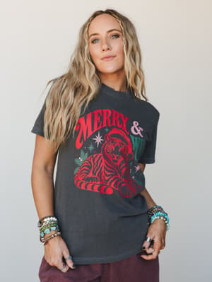 A washed charcoal graphic tee with a retro 'Merry & Wild' tiger-in-a-Santa-hat print, styled with stacked boho bracelets and dark burgundy bottoms — the kind of holiday outfit that actually sounds fun to wear.