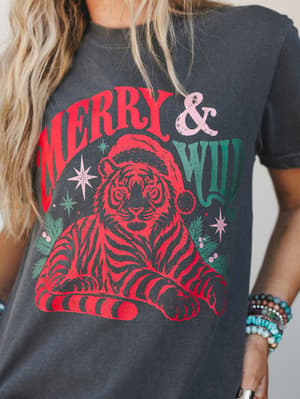 A close-up of a washed charcoal graphic tee featuring a retro 'Merry & Wild' tiger-in-a-Santa-hat print in red, green, and pink — worn with stacked beaded bracelets and a relaxed, effortless energy.