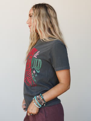 A side-profile shot of a model in a washed charcoal graphic tee with a bold 'Merry & Wild' tiger motif, styled with stacked beaded bracelets and deep plum bottoms — casual, expressive, and a little bit untamed.