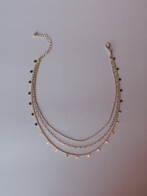 A delicate gold layered necklace with three mixed chains — curb, box, and paperclip — each strung with tiny flat disc charms, laid flat against a muted mauve-gray background.