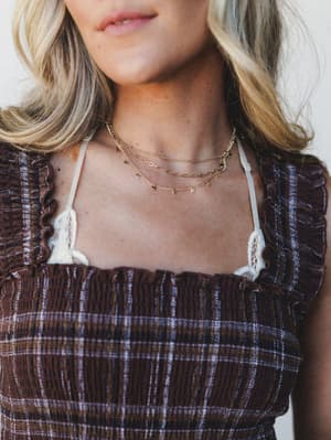 A close-up of layered gold chain necklaces styled against a smocked brown plaid top with ruffle trim — delicate coin charms catch the light in a way that feels collected, not coordinated.