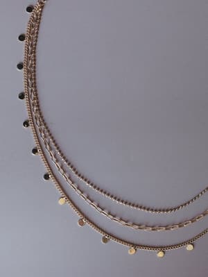 A close-up of a gold layered necklace fanned across a grey surface, featuring four distinct chain styles — curb, figaro, ball, and a coin-charm chain — that together have that effortlessly collected, already-been-wearing-it-for-years feel.