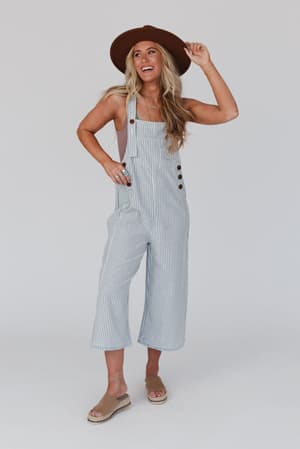 A smiling woman with long blonde hair wears the High Hopes Denim Overalls - Denim Stripe, paired with a wide-brimmed brown hat, mauve tank top, and tan sandals, standing against a plain light background.