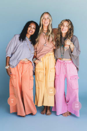 Three women stand barefoot before a light blue background, smiling and wearing Game On Cargo Wide Leg Pant - Black with casual tops. Height/size tags read: 5'4" XL, 5'8" Small, and 5'4" Small. The pants feature roomy cargo pockets.