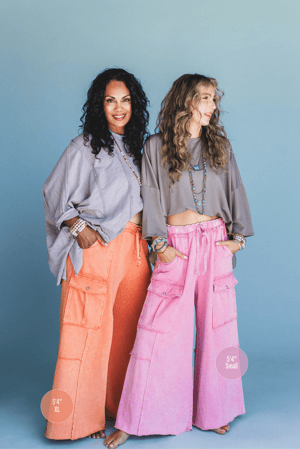 Two women stand side by side in front of a blue background, both wearing Game On Cargo Wide Leg Pant in Burnt Orange (sizes XL and Small), paired with loose tops and layered jewelry. Labels show both women are 5'4".