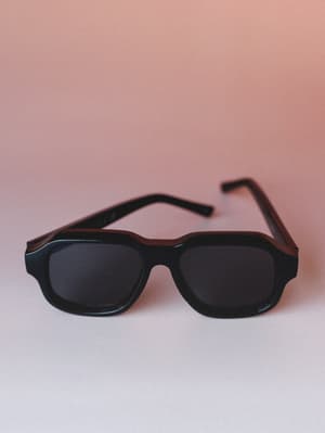 A pair of thick-framed black aviator-style sunglasses with dark lenses rests on a soft white surface against a warm blush-pink gradient background — the kind of shades that quietly pull the whole outfit together.