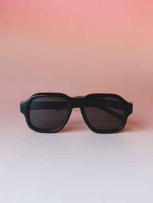 A pair of chunky black aviator-style sunglasses standing upright against a warm dusty rose gradient background — the kind of shades that make any outfit feel more intentional.