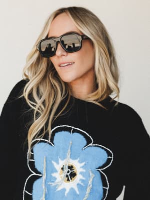 A relaxed, cool-girl close-up of a blonde model wearing black aviator-style sunglasses and a black graphic sweater with a large blue floral embroidered motif — effortlessly casual with a playful, artsy edge.