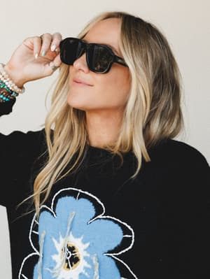 A sun-soaked close-up of a model in a black floral graphic sweater, holding oversized black sunglasses with that effortless 'just got back from somewhere good' energy — stacked bracelets, soft waves, the whole thing.