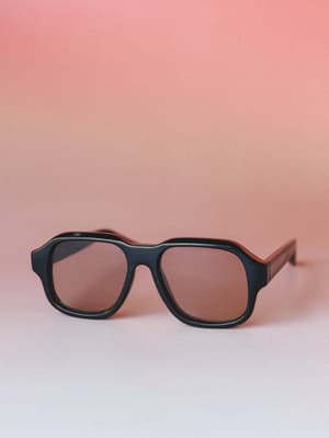 A pair of bold aviator-style sunglasses with a thick black frame and warm brown lenses, shot against a soft dusty rose gradient background — the kind of accessory that does half the work of an outfit without saying a word.