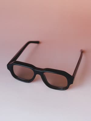 A pair of thick black-frame rectangular sunglasses with warm brown lenses rests against a soft dusty rose gradient backdrop — the kind of accessory that looks equally at home on a beach boardwalk or tossed on a café table.
