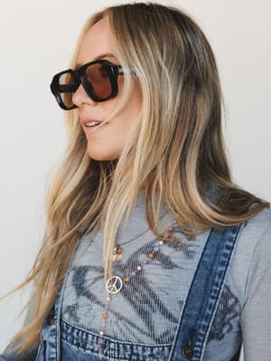 A close-up lifestyle shot of a woman in denim overalls and a grey top, layered with a peace sign pendant necklace and beaded chain — the oversized tortoise sunglasses are the star, shot in soft neutral light with an easy, sun-worn confidence.