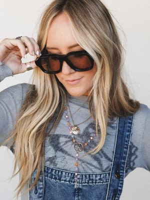 A model tilts her oversized black-frame sunglasses down with a soft smile, layered boho necklaces catching the light against a graphic grey top and denim overalls — the whole thing feels like a Saturday that turned out better than expected.
