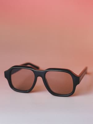 A pair of retro-inspired aviator sunglasses with a chunky black frame and warm brown lenses, shot up close against a dusty rose gradient backdrop — the kind of accessory that makes a simple outfit feel intentional.