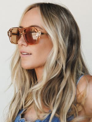 A close-up lifestyle shot of a model in oversized square-frame sunglasses with warm amber lenses, paired with a denim top — effortlessly cool with a sun-soaked, free-spirited feel.