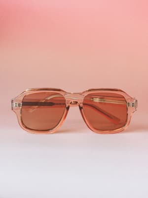 A pair of translucent tan aviator-style sunglasses with warm amber lenses, shot straight-on against a soft pink gradient backdrop — the kind of accessory that makes a Tuesday errand feel like a road trip.