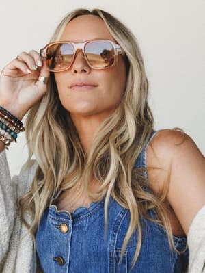 A sun-warmed close-up of a model in denim overalls and a cream cardigan, tilting oversized tan aviator-style sunglasses down with a stacked bracelet wrist — effortlessly cool, like she's about to say something worth listening to.