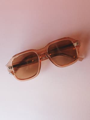 A pair of oversized square-frame sunglasses in a warm translucent tan, laid flat against a soft blush background — the kind of accessory that makes a coffee run feel intentional.