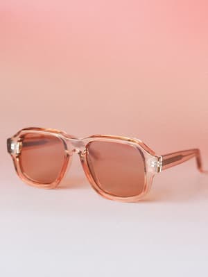 Translucent tan-framed square sunglasses with warm amber lenses, shot against a soft pink gradient backdrop — the kind of accessory that makes a Tuesday feel like a road trip.