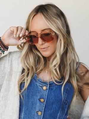 A sun-soaked close-up of a model tilting her amber-framed sunglasses down, layered in a button-front denim top and a cream knit cardigan, stacked bracelets and a pearl necklace finishing the look with that effortless lived-in energy.