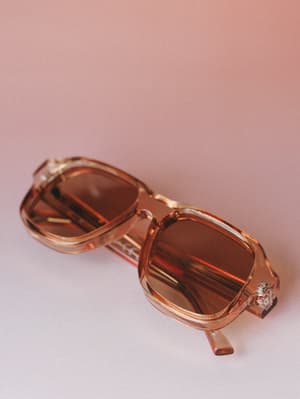 A pair of square-frame sunglasses with a clear amber-tan acetate frame and warm brown lenses, folded and resting against a soft dusty rose backdrop — the kind of sunnies that make a coffee run feel cinematic.