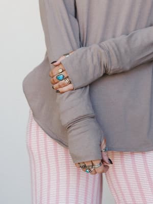 A close-up of crossed arms layered in stacked silver and turquoise rings, wearing a soft taupe long-sleeve top over a pink striped bottom — the kind of effortless, jewelry-heavy styling that feels personal and lived-in.