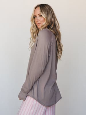 A relaxed long sleeve top in warm taupe-brown is shown from the back three-quarter angle, paired with pink striped bottoms — the kind of effortless, soft outfit that earns permanent residence on the bedroom floor (in a good way).