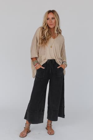 A woman wears the City Drifter Oversized Contrast Top in taupe with black wide-leg pants and tan sandals. She has long blonde hair, layered necklaces, colorful boho bracelets, and stands with her hands in her pockets against a plain background.