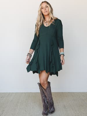 A long-sleeve mini dress in deep hunter green with a flowy ruffle front and handkerchief hem, styled with tall cowboy boots and layered boho jewelry for that effortless, earthy-cool vibe.