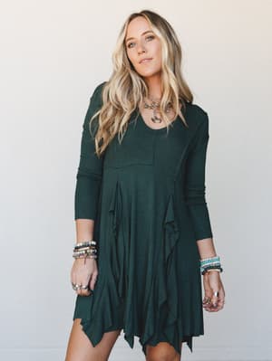 A flowy hunter green long-sleeve mini dress with ruffle and handkerchief hem details, styled with layered gold necklaces and stacked beaded bracelets against a soft neutral studio backdrop — easy, lived-in boho with just enough personality.