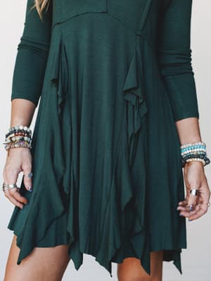 A close-cropped shot of a hunter green ribbed dress with flowing ruffle panels and a handkerchief hem, styled with stacked beaded bracelets and rings for that effortlessly collected, boho-everyday feel.