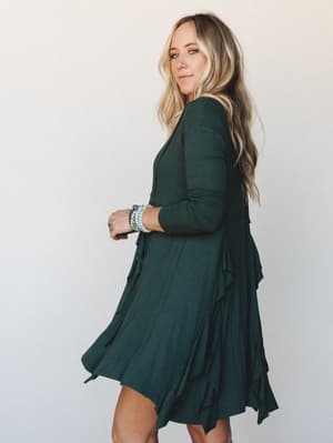 A model glances back over her shoulder in a hunter green long-sleeve dress with ruffle trim and a handkerchief hem — the kind of dress that does the work for you, stacked bracelets and all.
