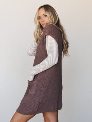 A longline bouclé knit sweater vest dress in deep mocha is layered over a fine-stripe long-sleeve top — the kind of effortless outfit that looks like you figured something out.