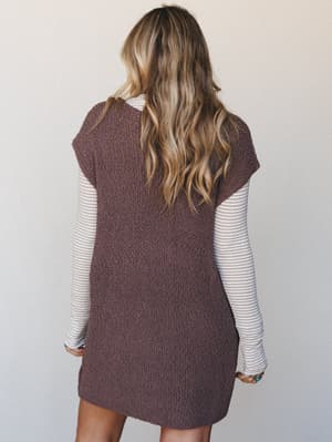 Shot from the back, a model layers a chunky knit sweater mini dress in mocha over a fine-stripe long-sleeve top — the kind of effortless, cozy-cool combo that makes fall dressing feel like a personality.