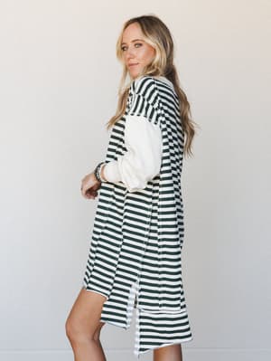 A striped knit mini dress in hunter green and cream worn over a cream long-sleeve top, shot in profile against a soft neutral studio wall — the kind of effortless layered look that feels just as right for a coffee run as it does for a casual dinner.