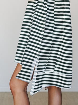 A close-up detail shot of a striped midi dress in hunter green and cream, showing the relaxed drape of the skirt, visible side slits, and exposed raw-edge seam stitching that gives it an intentionally casual, lived-in feel.