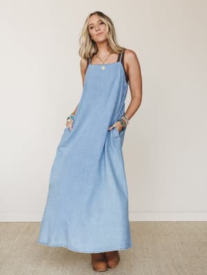 A flowy light-wash denim maxi dress styled with wood-soled clogs, stacked beaded bracelets, and a gold coin necklace — the kind of effortless outfit that looks like you tried just enough.