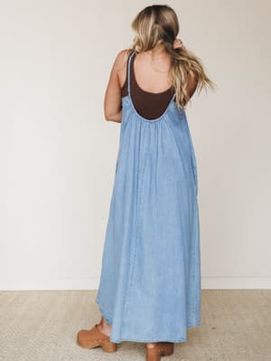 A breezy light denim maxi dress shot from behind, layered over a dark brown tank — the open back, gathered yoke, and easy silhouette give it that effortless 'grabbed it off the hook and just went' energy, grounded by tan wooden clogs.