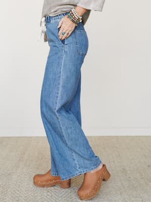 A side-angle shot of wide-leg medium wash denim jeans styled with a heathered gray top, wood-studded clogs, and layered beaded bracelets — the kind of outfit that does the work without trying too hard.