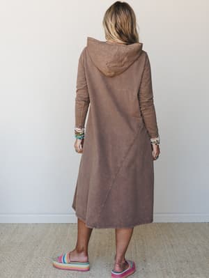 Shot from behind, a model wears a washed mocha midi hoodie dress with long sleeves and a diagonal seam detail — styled with stacked beaded bracelets and rainbow crochet platform sandals that somehow make the whole thing work.