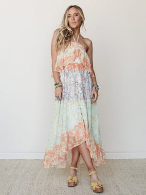 A flowy halter maxi dress in a dreamy patchwork of pastel floral prints — coral, mint, and lavender — styled with stacked boho bracelets and yellow platform sandals against a clean neutral wall.