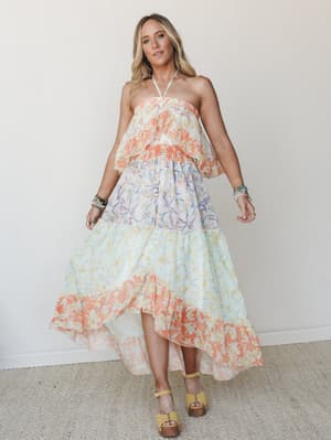 A flowy halter maxi dress in a color-blocked floral print moves through tiers of coral, lavender, and mint — styled with stacked bracelets and wood-platform sandals for a look that feels like a summer afternoon you don't want to end.