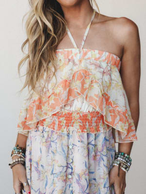 A sun-warmed floral dress with a halter neckline, layered ruffle bodice, and smocked waist — the kind of dress that looks equally at home on a beach boardwalk or a backyard birthday party, styled with stacks of beaded bracelets for that collected, we