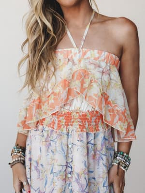 A sun-warmed floral dress with a halter neckline, layered ruffle bodice, and smocked waist — the kind of dress that looks equally at home on a beach boardwalk or a backyard birthday party, styled with stacks of beaded bracelets for that collected, we