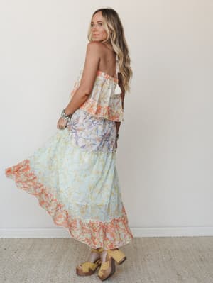A flowy maxi dress in a dreamy mix of coral and mint floral prints moves softly as the model glances back over her shoulder — the kind of dress that looks like it belongs somewhere warm, with clog sandals and stacked bracelets to match.