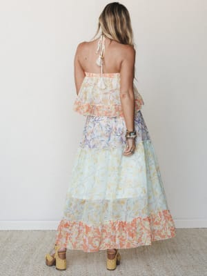 Shot from the back, a flowy tiered maxi dress in a color-blocked floral print moves through coral, soft lavender, and mint — styled with stacked boho bracelets and wood-platform sandals against a clean studio wall.