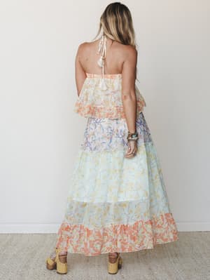 Shot from the back, a flowy tiered maxi dress in a color-blocked floral print moves through coral, soft lavender, and mint — styled with stacked boho bracelets and wood-platform sandals against a clean studio wall.